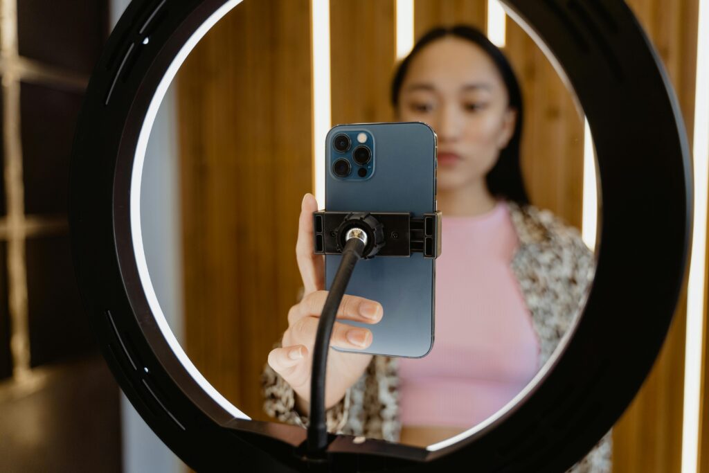 Young woman using a smartphone held by a ring light for video recording indoors.